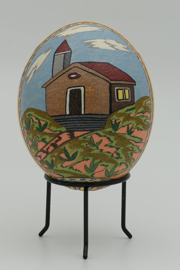 Intricately designed decorative egg featuring a painted house on a hill, set against a vibrant blue sky.