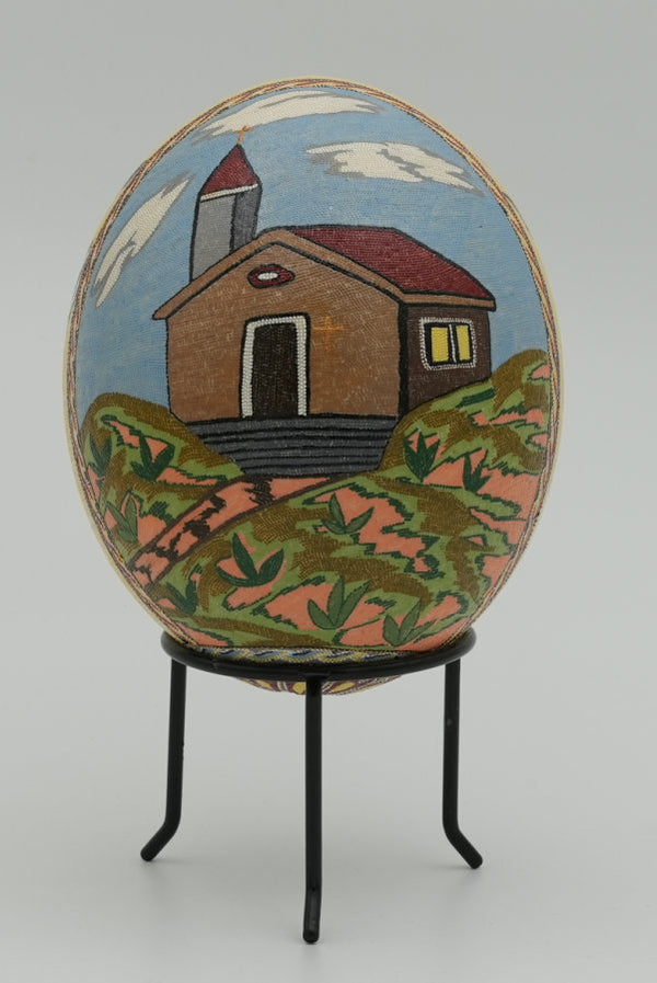 Intricately designed decorative egg featuring a painted house on a hill, set against a vibrant blue sky.