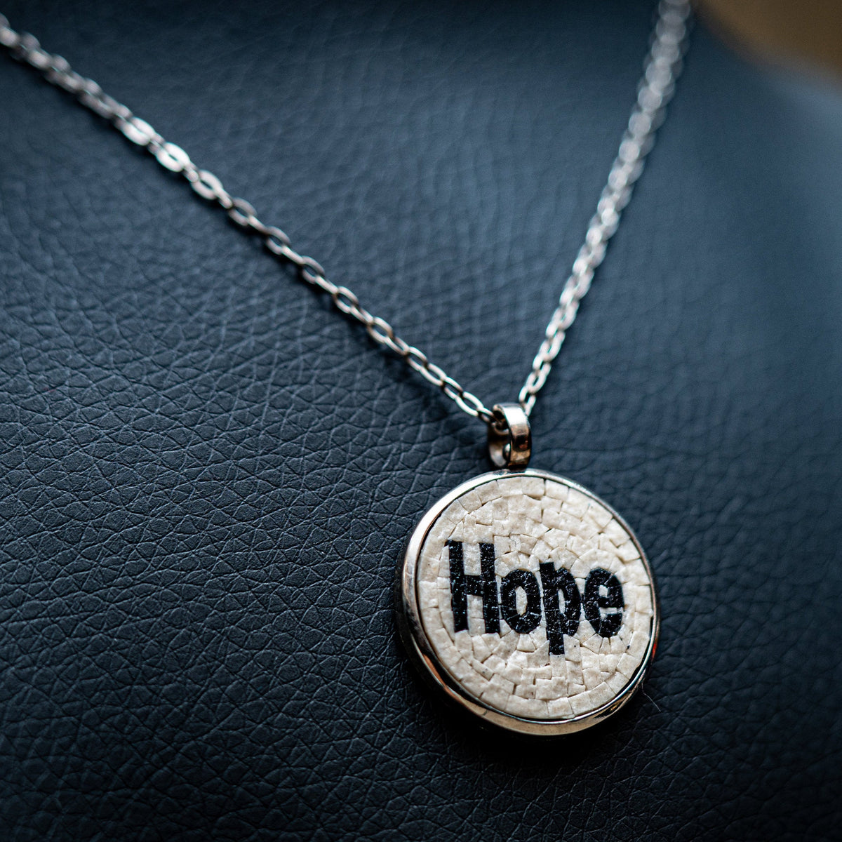 Silver pendant necklace featuring a textured round charm with the word 'Hope' elegantly displayed on a dark leather background