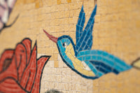 Vibrant mosaic artwork featuring a blue hummingbird with yellow accents perched beside a red flower on textured tile.