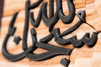 Intricate carved wooden wall art featuring elegant Arabic calligraphy on a textured sandstone background.